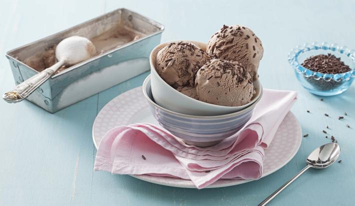 Fotografia em tons de azul e rosa em uma bancada de madeira azul clara, um pano rosa, um pratinho rosa redondo com potinhos de porcelana em cima e as bolas de sorvete de brigadeiro com granulado para decorar. Ao lado, um potinho com granulados e colher.