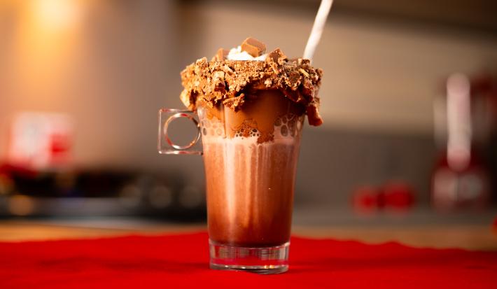 Fotografia em tons de vermelho com um copo alto ao centro. Dentro do prato existe uma bebida de café com chocolate. As bordas do copo estão cobertas por pasta de KITKAT e por cima existe vários pedaços de KITKAT triturados.