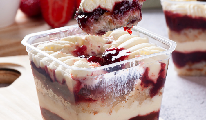 Fotografia mostra um bolo recipiente transparente mostrando camadas de um bolo de pote com creme branco e geléia de frutas vermelhas.
