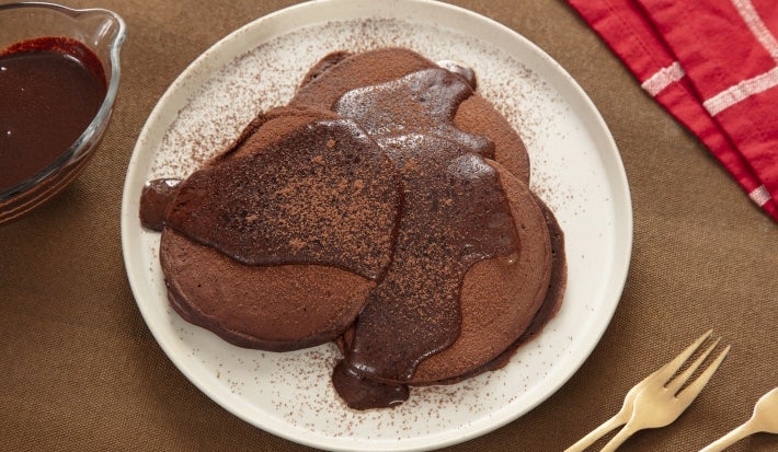 Fotografia de um prato de vidro grande e raso, branco, ao lado de alguns garfos dourados, um pano vermelho e branco e um potinho de vidro com calda de chocolate. Sobre o prato tem três panquecas de nescau com uma calda de nescau extra cacau, também. Fotografia de um prato de vidro grande e raso, branco, ao lado de alguns garfos dourados, um pano vermelho e branco e um potinho de vidro com calda de chocolate. Sobre o prato tem três panquecas de nescau com uma calda de nescau extra cacau, também.