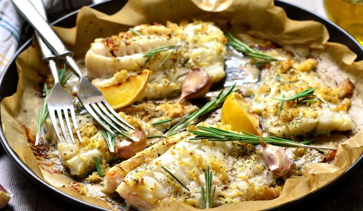 Fotografia de quatro filés de peixe assados com MAGGI MEU SEGREDO 7 Vegetais, ervas frescas e amêndoas. O assado está sobre papel manteiga e dentro de uma forma preta.