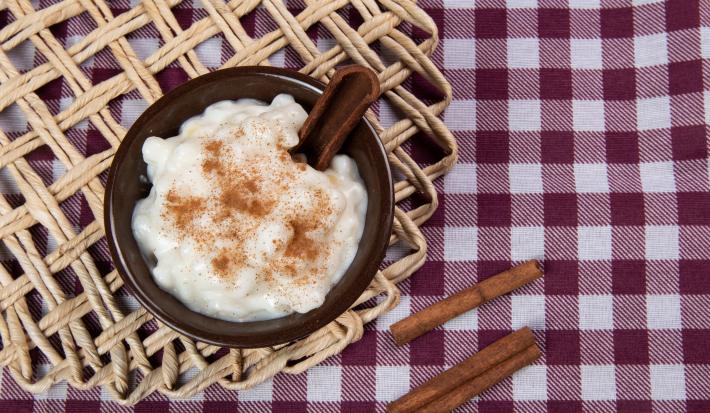 Fotografia vista de cima de um recipiente pequeno de vidro marrom com uma receita de canjica vegana. O recipiente está sobre um pano de mesa quadriculado de tom vinho e branco, ao lado do recipiente tem três paus de canela.