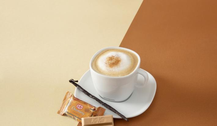 Foto da receita de cappuccino vanilla servida em uma xícara branca com um kit kat churros ao lado em fundo marrom claro e escuro