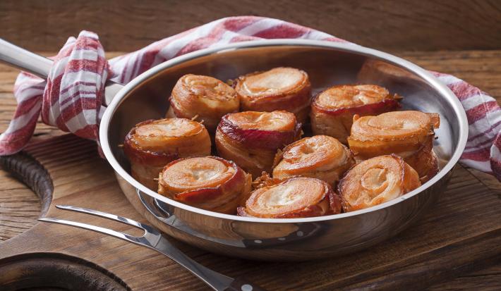 Fotografia em tons de marrom, prata e vermelho de uma bancada de madeira e uma tábua de madeira, sobre ela um panela de inox com medalhões de frango com bacon. Ao lado um garfo de servir, ao fundo um paninho branco e vermelho.