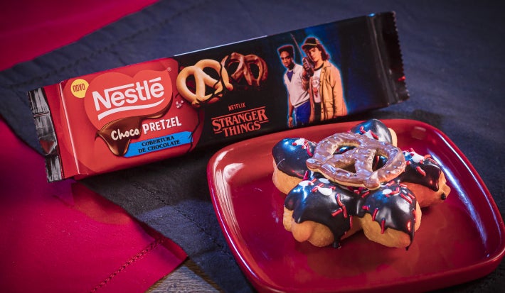 Donut em formato de flor, coberto com chocolate escuro e branco, detalhes vermelhos e um ChocoPretzel crocante no centro.