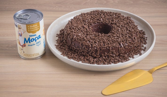 Foto da receita de Brigadeirão Moça, servido em um prato branco, decorado com muitos granulados de chocolate. O prato está entre uma lata de Leite Moça e uma espátula dourada, tudo sobre uma bancada de madeira