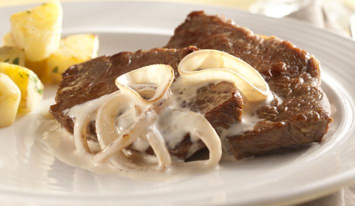 foto em tons de branco e marrom de uma bancada vista de cima. Contém um prato branco redondo com um bife com cebolas com creme e abaixo um pano branco.
