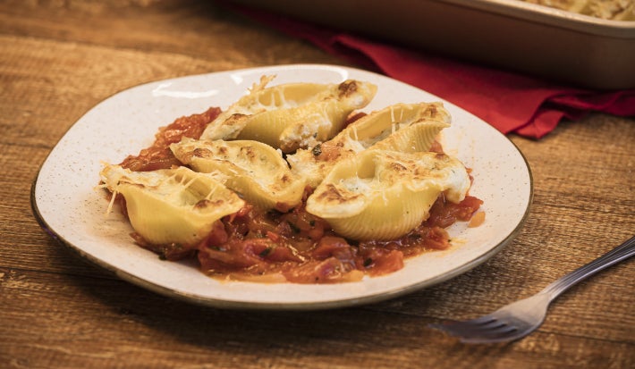 Foto da receita de Conchiglione recheado com 3 queijos Tirolez, servidos lado a lado em um prato branco. Eles estão recheados e cobertos com mais queijo, sobre um molho rústico de tomate