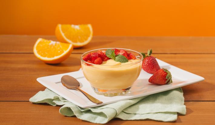 Fotografia em tons de laranja em uma bancada de madeira, um fundo laranja, um recipiente branco retangular, um pano verde claro e uma tacinha de vidro com a mousse de laranja dentro dela, com morangos para decorar. Ao fundo, uma laranja partida ao meio.