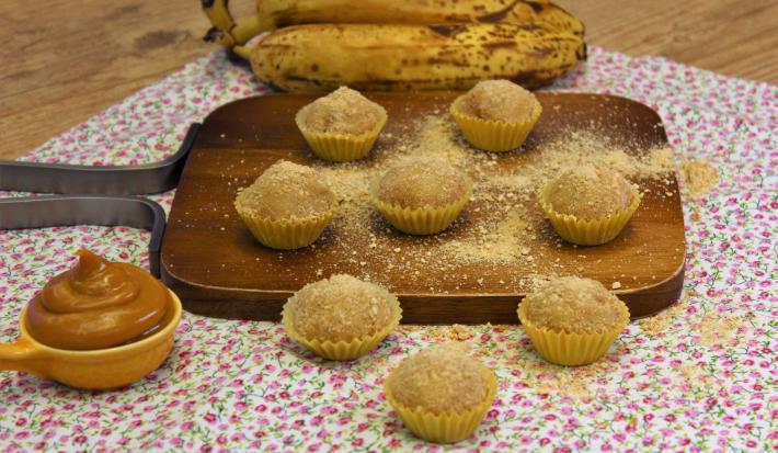 Fotografia em tons de amarelo em uma bancada de madeira de cor marrom. Ao centro, uma toalha branca florida e em cima, há uma tábua de madeira contendo os brigadeiros. Ao fundo, há um cacho de bananas.