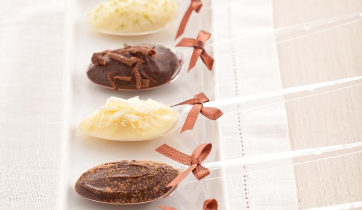 fotografia em tons de branco e marrom tirada de um recipiente retangular com 5 colheres descartáveis e todas contém uma trufa nos sabores chocolate e chocolate branco. Nas colheres um laço para decorar.