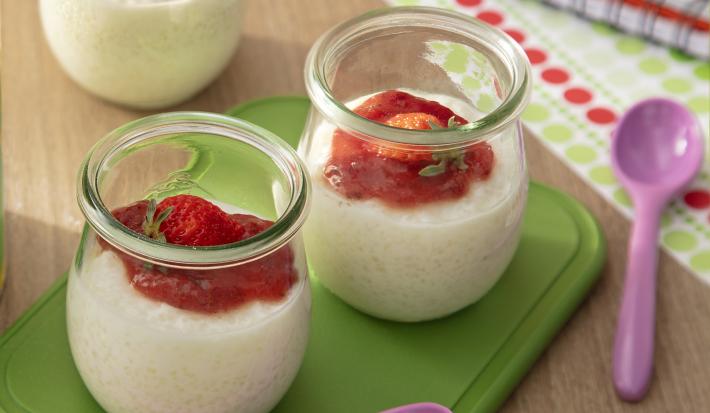 Fotografia de três recipientes de vidro pequenos com pudim de tapioca de ninho um creme de morango por cima. Dois desses recipientes estão sobre um apoio verde retangular, ao lado de uma colher roxa pequena.