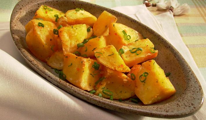 Fotografia em tons de amarelo em uma mesa com uma toalha quadriculada em amarelo, laranja e verde e um recipiente oval fundo com batatas cozidas e cebolinha-verde dentro dele.