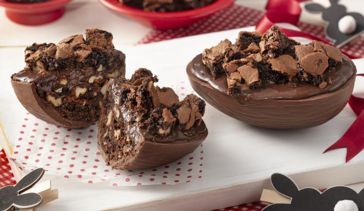 Fotografia em tons de branco e vermelho de uma bancada branca com uma tábua branca e uma paninho branco com bolinhas vermelhas, sobre ela dois ovos de pascoa recheados. Ao lado coelinhos. Ao fundo dois recipientes vermelhos com brownie e chocolate.