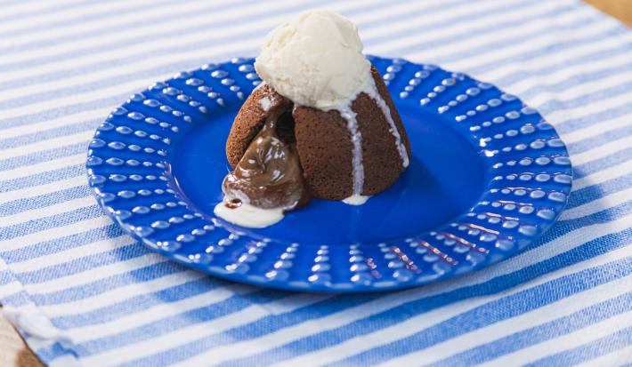 Fotografia em tons claros de um prato azul escuro raso apoiado em um pano azul claro com listras brancas. Sobre o prato tem um petit gateau aberto com a calda saindo, e uma bola de sorvete de creme escorrendo sobre a sobremesa.