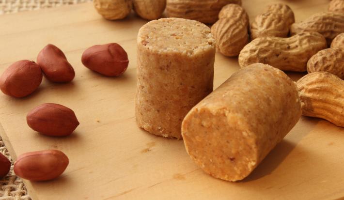 Fotografia em tons de caramelo com uma paçoca de formato cilíndrico ao centro. Em volta da paçoca existe alguns amendoins