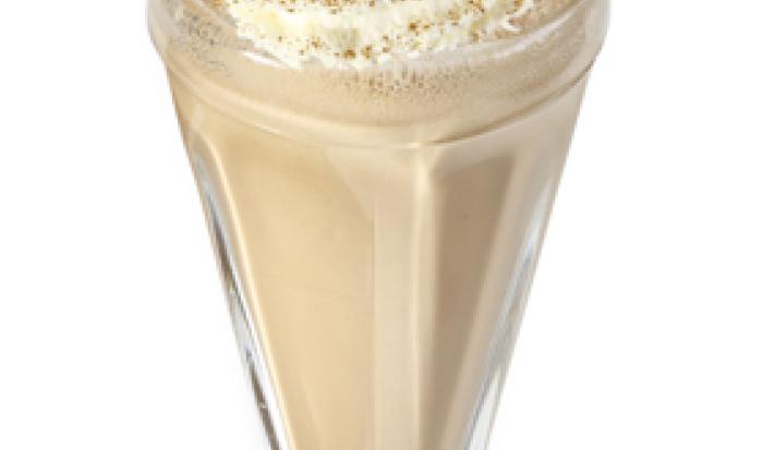 Fotografia vista de frente em tons de branco e marrom, ao centro um copo transparente com milkshake de café dentro e creme e canela por cima para decorar.