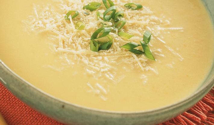 Fotografia em tons de creme e vermelho, com refratário cinza ao centro contendo um creme amarelo decorado com queijo e cebolinha picada sobre guardanapo vermelho, com colher ao lado e tudo sobre aparador de palha na cor bege. Fotografia em tons de creme e vermelho, com refratário cinza ao centro contendo um creme amarelo decorado com queijo e cebolinha picada sobre guardanapo vermelho, com colher ao lado e tudo sobre aparador de palha na cor bege.