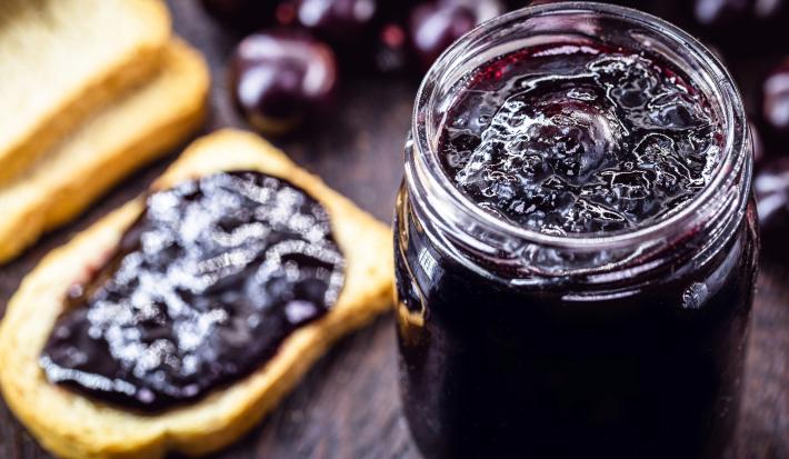 Fotografia de um recipiente de vidro com geleia de jabuticaba sobre uma mesa de madeira escura. Ao lado do recipiente tem algumas torradas, e uma delas está com um pouco de geleia, ao lado também de algumas jabuticabas.