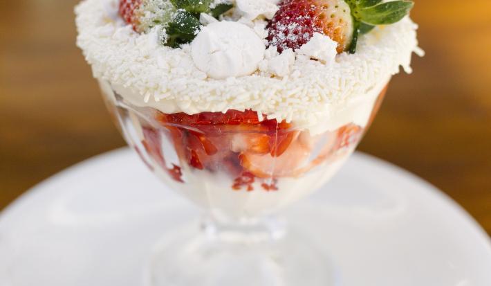 Fotografia de uma taça de sobremesa com merengue, morangos, suspiro e granulado branco nas bordas. O recipiente está em cima de um prato branco de sobremesa, com uma colher pequena. O prato está sobre uma mesa marrom.