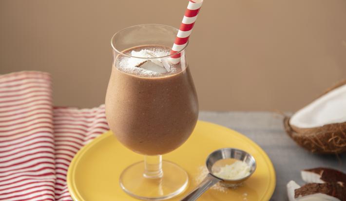 Fotografia em tons de marrom em uma bancada de madeira de cor cinza. Ao centro, um prato amarelo contendo uma taça com o smoothie. Ao fundo, há um pano branco listrado de vermelho e um coco partido ao meio.