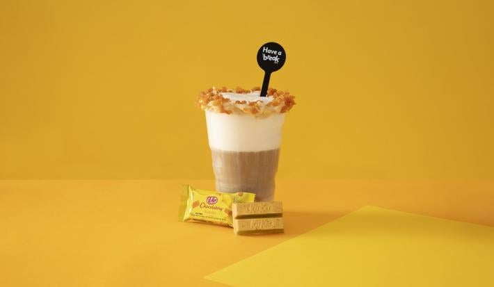 Foto em tons de amarelo da receita de latte salted caramel servida em um copo grande de vidro com um KIT KAT banana ao lado em fundo amarelo
