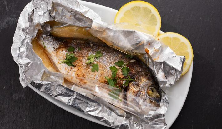 Fotografia em tons de cinza em uma bancada de madeira de cor preta. Ao centro, um prato branco contendo o peixe em um papel alumínio com duas rodelas de limão ao lado.
