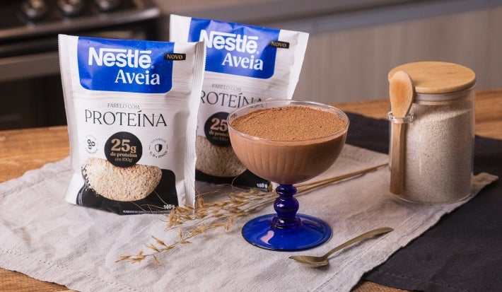 Foto da receita de mousse proteico de chocolate servida em uma taça de vidro sobre um pano neutro em cima de uma mesa de madeira com um pote de aveia ao lado com uma colher de madeira acoplada