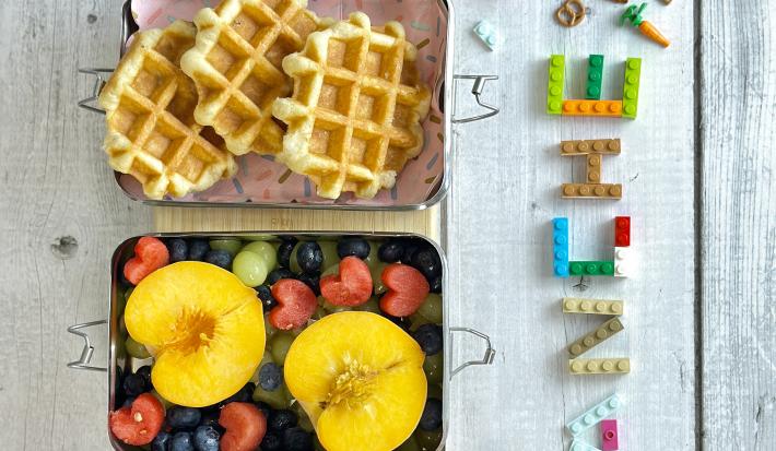 Fotografia em tons coloridos com uma lancheira com waffles e frutas dentro dela. Ao lado letras de brinquedo escrito a palara lanche em uma bancada azul em madeira.