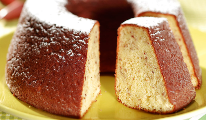 Fotografia em tons de verde em uma mesa de madeira com uma toalha verde quadriculada. Ao centro, um suporte para bolo verde com o bolo de pinhão em cima dele.