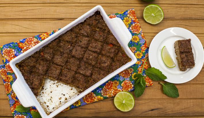 Fotografia em tons de marrom em uma mesa de madeira com um pano florido, um refratário com alças laterais brancas com o quibe de carne dentro dele. Ao lado, um pratinho branco com uma fatia do kibe e rodelas de limão.