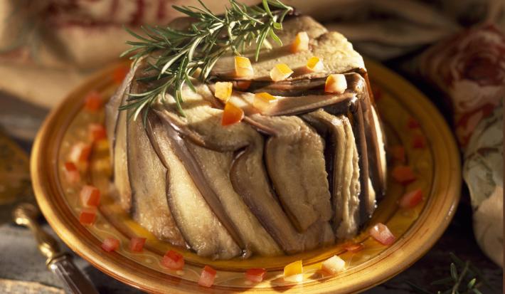 Fotografia em tons escuros de um bolo de berinjela, com cubinhos de tomate e alecrim por cima, em um prato amarelo. Ao lado, um ramo de alecrim, e ao fundo, um pano decorado, que está sobre uma mesa de madeira em tom escuro.