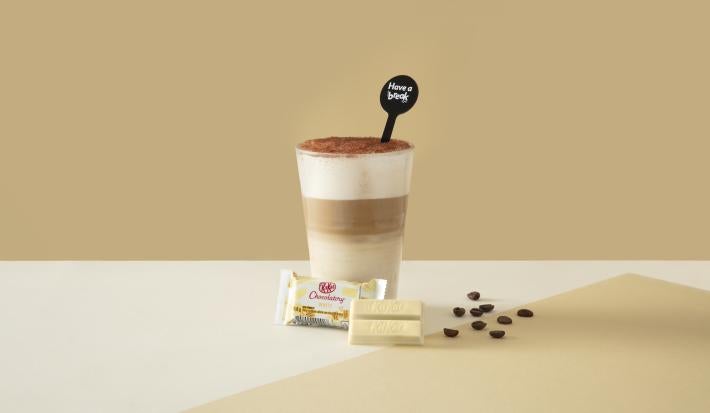Foto da receita de Latte Tiramisú. Observa-se um fundo bege com o copo ao centro, dividido em 3 fases: embaixo, uma mais clara, a do meio, mais escura - de café, e a de cima, com creme. Por fim, chocolate polvilhado. À frente, um KITKAT de chocolate branc