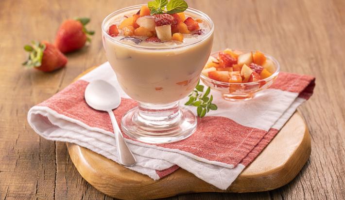 Foto em tons de vermelho da receita de taça cremosa de frutas servida em uma taça de vidro sobre um pano listrado com uma colher prateada e uma taça menor com pedaços de frutas ao lado. Tudo sobre uma tábua de madeira e há morangos ao fundo