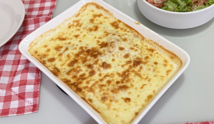 Foto da receita de tilápia gratina ao molho branco e creme de cebola, servida em uma travessa retangular branca, com um aspecto gratinado apetitoso, tudo em uma mesa com uma toalha xadrez vermelha e branca além de outro recipiente com uma salada dentro
