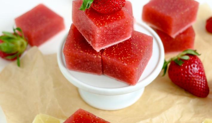 Fotografia em tons de vermelho em uma bancada de madeira com um pano bege, um suporto de bolo branco, com balas de goma de morango. Ao lado, morangos e fatias de limão siciliano.