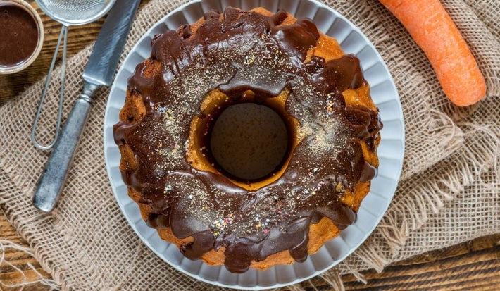 Foto vista de cima de um bolo de cenoura com cobertura de chocolate e confeitos coloridos, sobre um prato branco. O bolo está apoiado em um tecido rústico de juta, com utensílios ao redor e uma cenoura ao lado, remetendo aos ingredientes da receita.