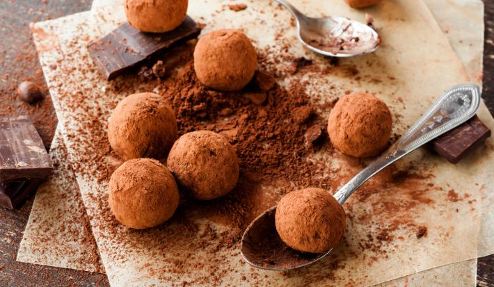 Fotografia vista de cima de algumas trufas de café e chocolate passadas no cacau em pó, sobre uma tábua de madeira salpicada com cacau em pó. Tem alguns pedaços de chocolate em volta das trufas e uma colher segurando uma delas.