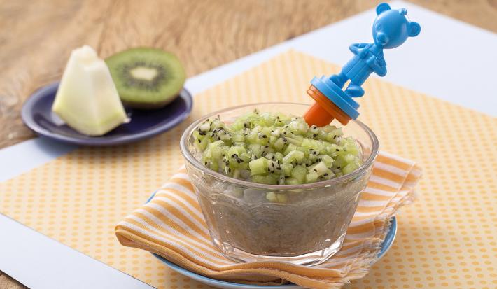 Fotografia de uma bancada de madeira com paninho laranja com bolinhas, um potinho transparente com uma papinha e cubinhos de kiwi e uma colher de ursinho azul. Ao fundo um pratinho com um pedaço de melão e um pedaço de kiwi.