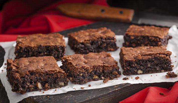 Foto de 6 pedaços iguais de brownie em cima de uma toalha branca e com uma faca de serra ao fundo da foto