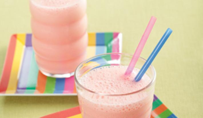 Fotografia em tons de rosa em uma bancada de madeira bege com dois pratos quadrados com listras coloridas e dois copos de vidro com a vitamina de Nesquik dentro e dois canudinhos, um azul e outro rosa dentro de cada copo.