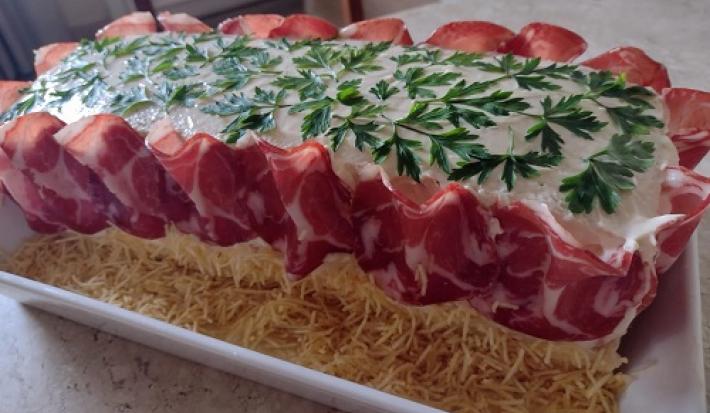 Foto da receita de pão surpresa decorado com folhas verdes por cima e presunto de parma cobrindo os lados, além de batatas palhas ao fundo da travessa