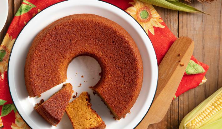 Bolo de milho feito com Farinha Láctea Nestlé em um prato branco, com dois pedaços cortados e espigas de milho em volta.