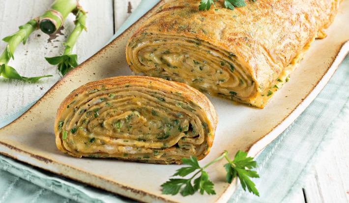 Fotografia em tons de verde em uma bancada de madeira clara com um recipiente retangular bem comprido e grande com a omelete em forma de rocambole.
