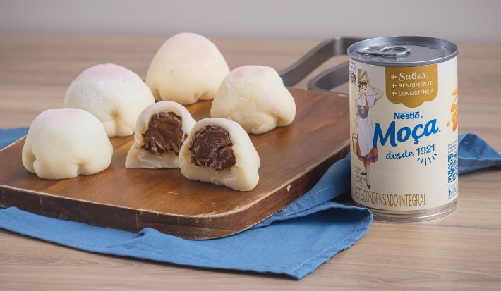 Receita de Mochi de Brigadeiro, servida em pequenas bolinhas brancas, recheadas com brigadeiro enrolado, sobre uma tábua de madeira e um tecido na cor azul ao lado de uma Lata de Leite Moça