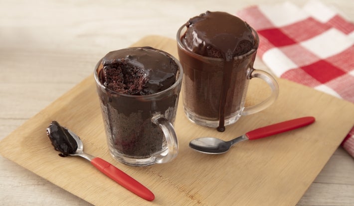 Foto da receita de bolo de chocolate de caneca feito com nescau extra cacau servida em duas canecas de vidro acompanhadas de duas colheres de cabo laranja sobre uma tábua de madeira. Esses elementos sobre uma mesa de madeira com um pano quadriculado