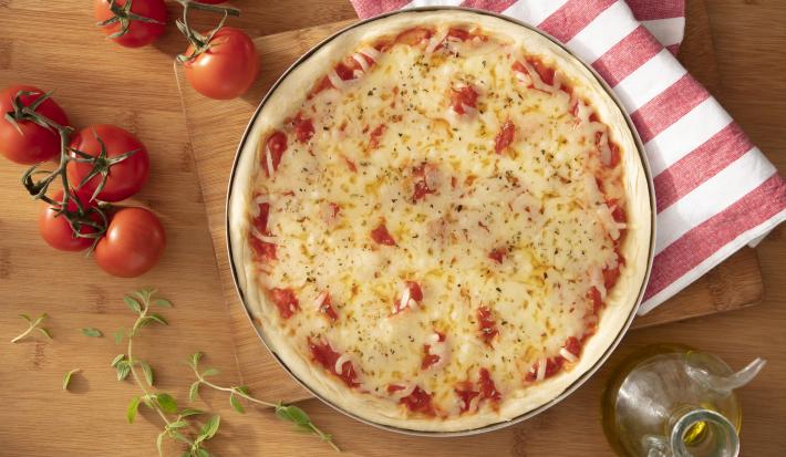 Imagem vista de cima de uma pizza redonda de queijo muçarela. Ao lado, um paninho listrado branco e vermelho e tomates.