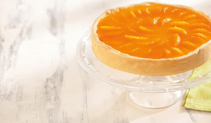 foto em tons de branco e laranja de uma bancada branca vista de cima. Contém um suporte para servir sobremesas transparente com uma torta com cobertura de tangerina por cima.
