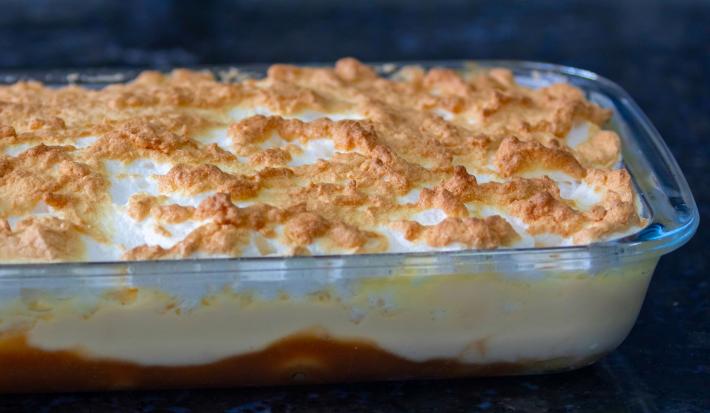 Imagem de uma travessa de vidro com a sobremesa chico balanceado. É possível ver a camada de caramelo com banana, creme branco e merengue dourado.