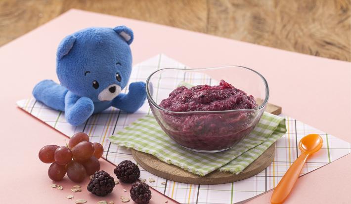 Fotografia de uma bancada de madeira com um paninho rosa, sobre ele um potinho transparente com uma papinha roxa, à frente uvas e amoras e ao lado um ursinho de pelúcia azul.